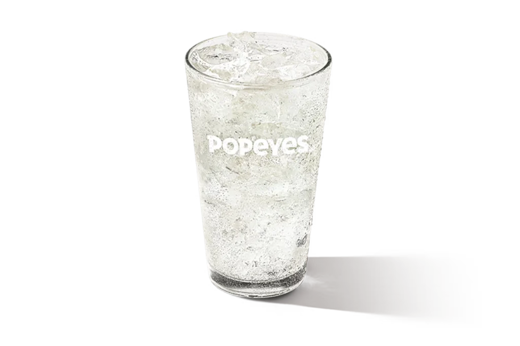 "Glass of seltzer water with ice cubes, Popeyes logo on the glass, on a white background."